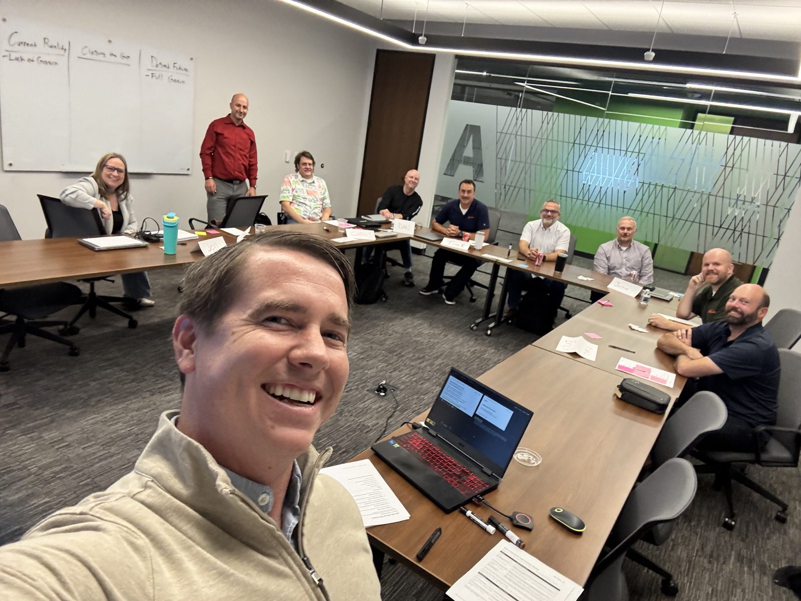 Shem Hatfield taking a selfie with a group of workshop participants seated around conference tables in Waukesha, Wisconsin, with a current-reality and desired-future whiteboard exercise behind them